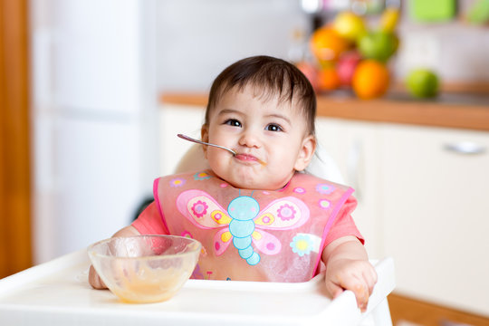 Baby girl eating with spoon and sitting in a high chair for feeding