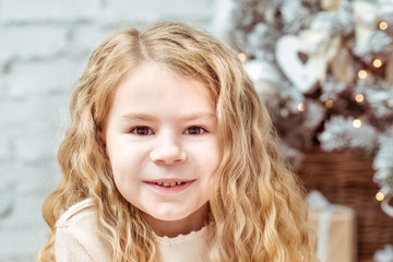Fototapeta premium Pretty blond little girl sitting with long curly hair under the Christmas tree and smiling, closeup portrait