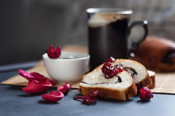 Slices of butter poppy roll, served with cherry jam. Large ceramic cup with hot drink. Black table and bamboo mat. Red dry flowers are scattered around. The delicious steam comes from mug