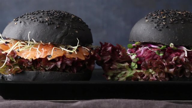 Two homemade black burgers with salted salmon, beetroot cutlet, sprouts and green salad on black plate over dark background