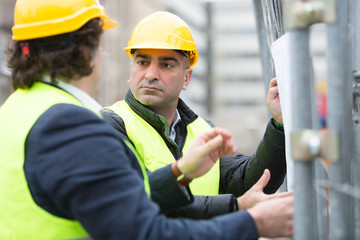 Two architects wearing hardhat and yellow jacket reading office blueprints against scaffolding at construction site