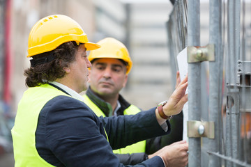 Two engineers wearing hardhat and yellow jacket reading office blueprints against scaffolding at construction site
