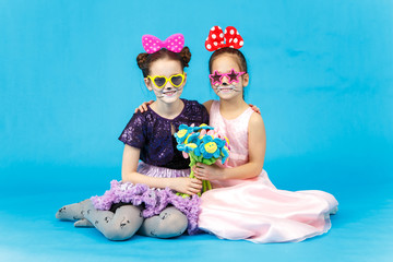 Two smiling girls in funny sunglasses sitting on blue background