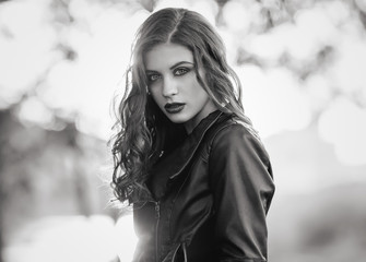 Black and white portrait of young beautiful woman in leather jacket with wavy hairstyle on a blurred forest background