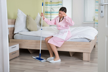 Tired Housekeeper Sitting On Bed With Mop