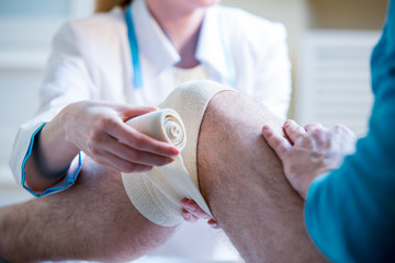 Female doctor is rewinding knee bandage to man. Doctor taking care of a patient with trauma of...