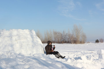The man sitting near an igloo and drinking tea from thermos  in the winter
