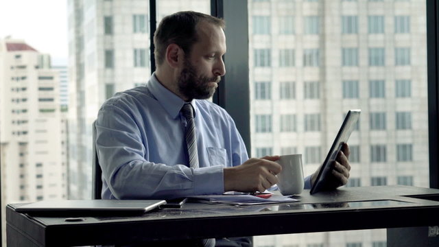 Young businessman reading something on tablet and drinking coffee in office
