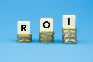 Word ROI (Return of Investment) on stacks of gold coins over blue background