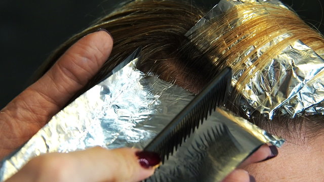 Hairdresser hands folding foil underlay with dyed locks of hair of beautiful young woman in a hair and beauty salon. Highlighting or lowlighting hair, professional hair coloring process.  
