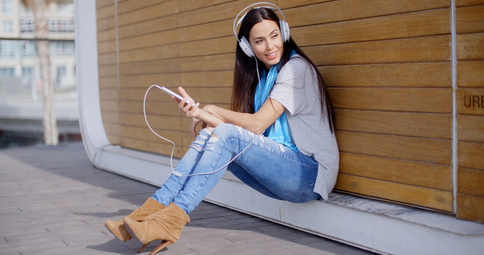 Pretty trendy young woman enjoying her music on her mobile phone smiling as she relaxes on the ground in front of a closed urban kiosk