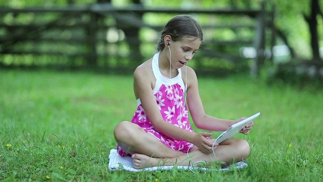 Beautiful girl in pink dress with tablet computer sits on green grass in garden. Little girl with tablet pc sits on the grass in garden. Girl communicates through Skype. Female with tablet computer