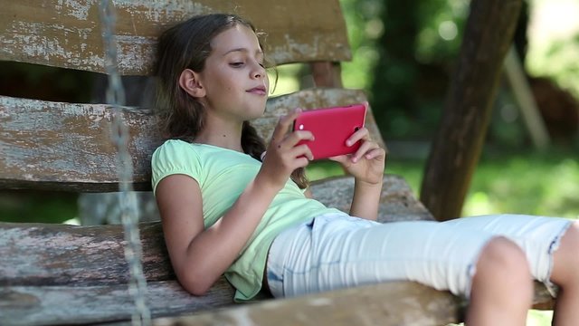 Girl with red smartphone sits on a swing bench in the garden and listens to music. Attractive girl uses smartphone. Beautiful girl with red smartphone sits on the swing bench