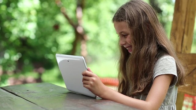 Beautiful girl with white tablet computer sits at the table. Girl plays the game on his Tablet PC. Little girl with tablet computer sits on the bench in garden. Female with tablet computer