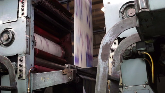 Handheld shot of the cyan printing drum and the uncut newspaper being printed running through an industrial offset newspaper printing press.