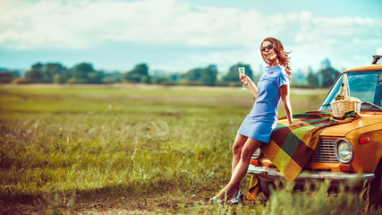 Beautiful woman on the picnic is drinking wine near a car. - obrazy, fototapety, plakaty