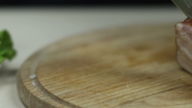 Кухонный нож режет бекон на разделочной доске из дерева.
