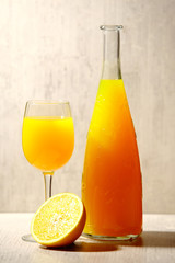 Fresh orange lemonade in a glass next to the whole fruit and orange slices on a light gray background. Selective focus