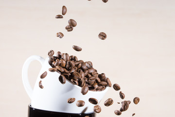 Close up of white cup standing on black cup with falling down brown roasted coffee beans.