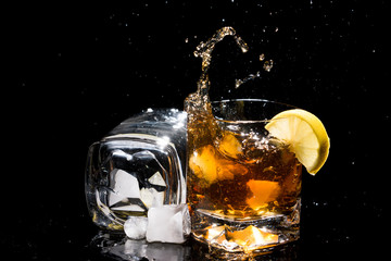Two high quality glasses of whiskey, one lies on side with two pieces of ice near  and other with falling ice and lemon with splashes.