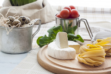 Raw homemade pasta, camembert, quail easter eggs in a aluminum cup, green lettuce, tomatoes and flour on wooden table