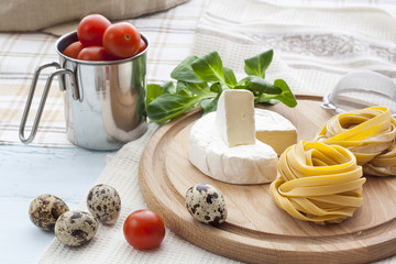 Raw homemade pasta, camembert, quail easter eggs in a aluminum cup, green lettuce, tomatoes and flour on wooden table