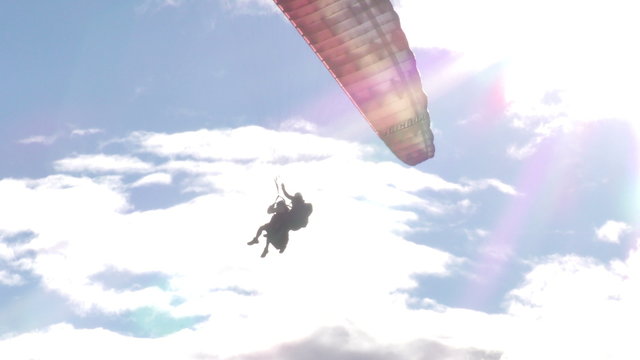 A paraglider glides through a partly cloudy sky silhouetted against bright sunlight with lens flares near Banos, Ecuador, the camera following from a low angle across the expansive sky.