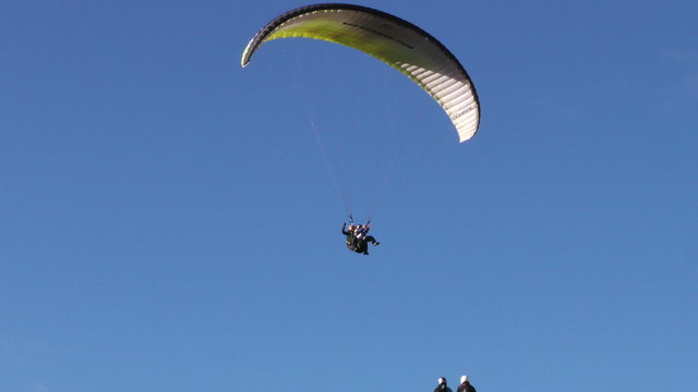 A paraglider descends and lands on a grassy hill under a clear blue sky as onlookers watch from the ground near Banos, Ecuador, the full motion from mid-air to touchdown in a wide low-angle view.