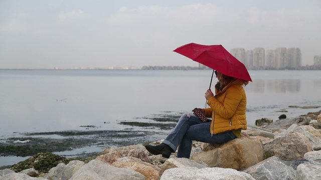 Woman with umbrella sitting alone in rocks on the seaside bank and be lonely, rainly day