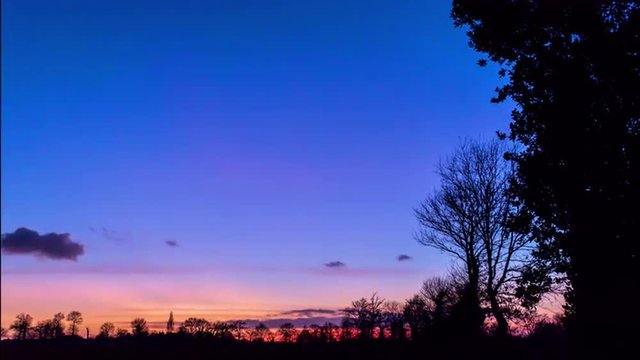 timelapse of a cloudy sunset with a beautiful orange and blue sky behind trees a the edge of a deserted field during winter