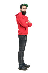 Side view of young bearded man in red hoodie wearing baseball cap with crossed arms. Full body length portrait isolated over white studio background. 