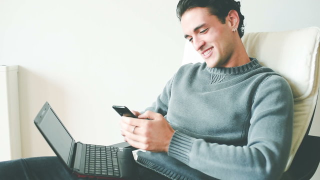 Young handsome caucasian brown hair man sitting on the sofa laptotp on his knee holding a smartphone tapping the screen, shaking his head, smiling - communication, happiness, social network concept