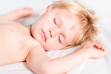 Obraz na płótnie Canvas Close-up portrait of handsome toddler boy with blond hair sleeping on white bed. Carefree childhood concept
