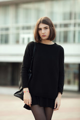 Street portrait of young elegant brunette hipster woman in black blouse and skirt leaning against a blurred city background