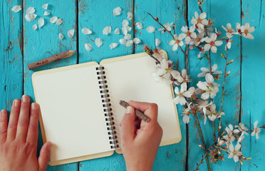 woman writing on blank notebook next to spring white cherry blossoms tree on vintage wooden table. 