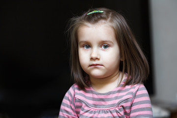 Close-up portrait of a serious little girl against dark background. Child looks into the camera.