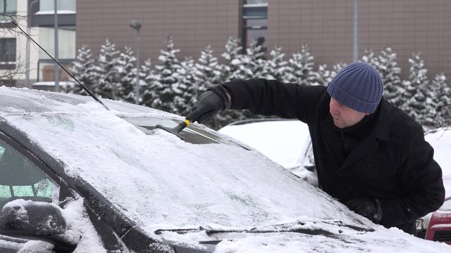 Man with scraper scraping snow and ice from car front window windscreen in living house district in winter season. Static shot. 4K
