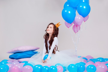 Cute little princess girl sitting among balloons in room over white background. Looking at camera....