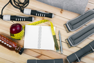 Dumbbells, Jump Rope, Towel, Ankle Weights, Tape Measure, Apple, Bottle Of Juice, Notepad To Workout Plan On Wooden Floor. Sport Fitness Background