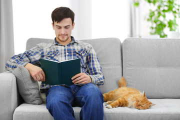 Reading young man with fluffy rad cat on a sofa