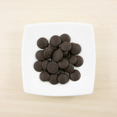 The group of dark chocolate buttons and small white square disk on light brown wooden board.