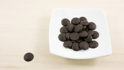 The group of dark chocolate buttons and small white square disk on light brown wooden board.