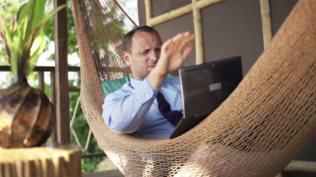 Angry businessman destroying laptop while lying on the hammock on terrace
