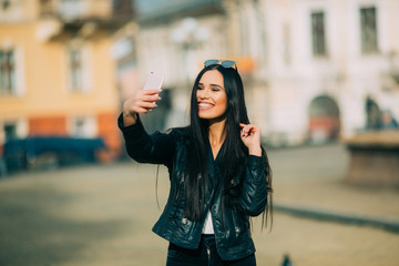 Young beautiful casual girl takes a selfie on her cell phone with sunglasses in jacket an black bag smile to camera on  spring city street. Spring outfit, Spring street look.