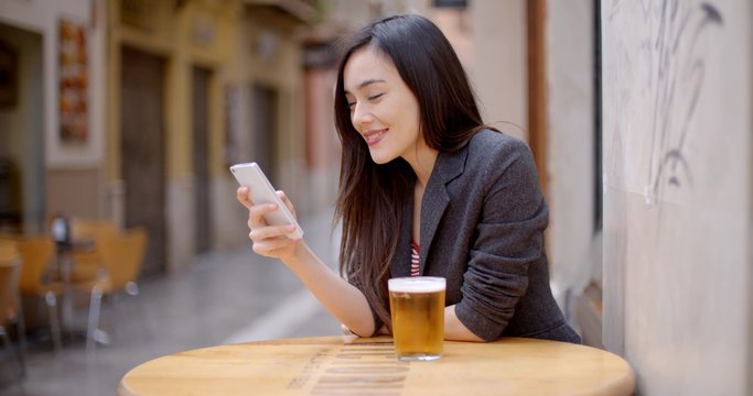 Smiling young woman relaxing with a beer at an open-air table in town with her mobile phone in her hand as she reads a text message.