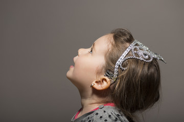 Lovely expressive little girl wearing silver princess crown looking up, studio shot on gray...