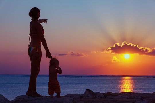 Black Silhouette Of Happy Family Walking On Sea Beach - Mother Hold Baby Son Hand, Point Sun And Look At Sunset Sky. Active Parent Lifestyle, Outdoor Activity On Tropical Summer Vacation With Children