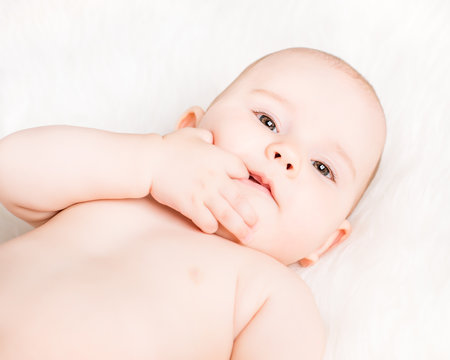 Closeup portrait of a cute smiling baby lying on white fur and sucking fingers