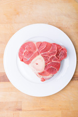 one piece of raw beef meat with the bone in the middle on a white plate with a background kitchen wooden board