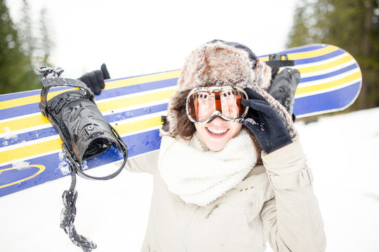Snowboarding.Young beautiful woman with ski mask holding her snowboard at ski slope Young woman in ski resort holding snowboard on her shoulders and smiling.Concept of winter holiday